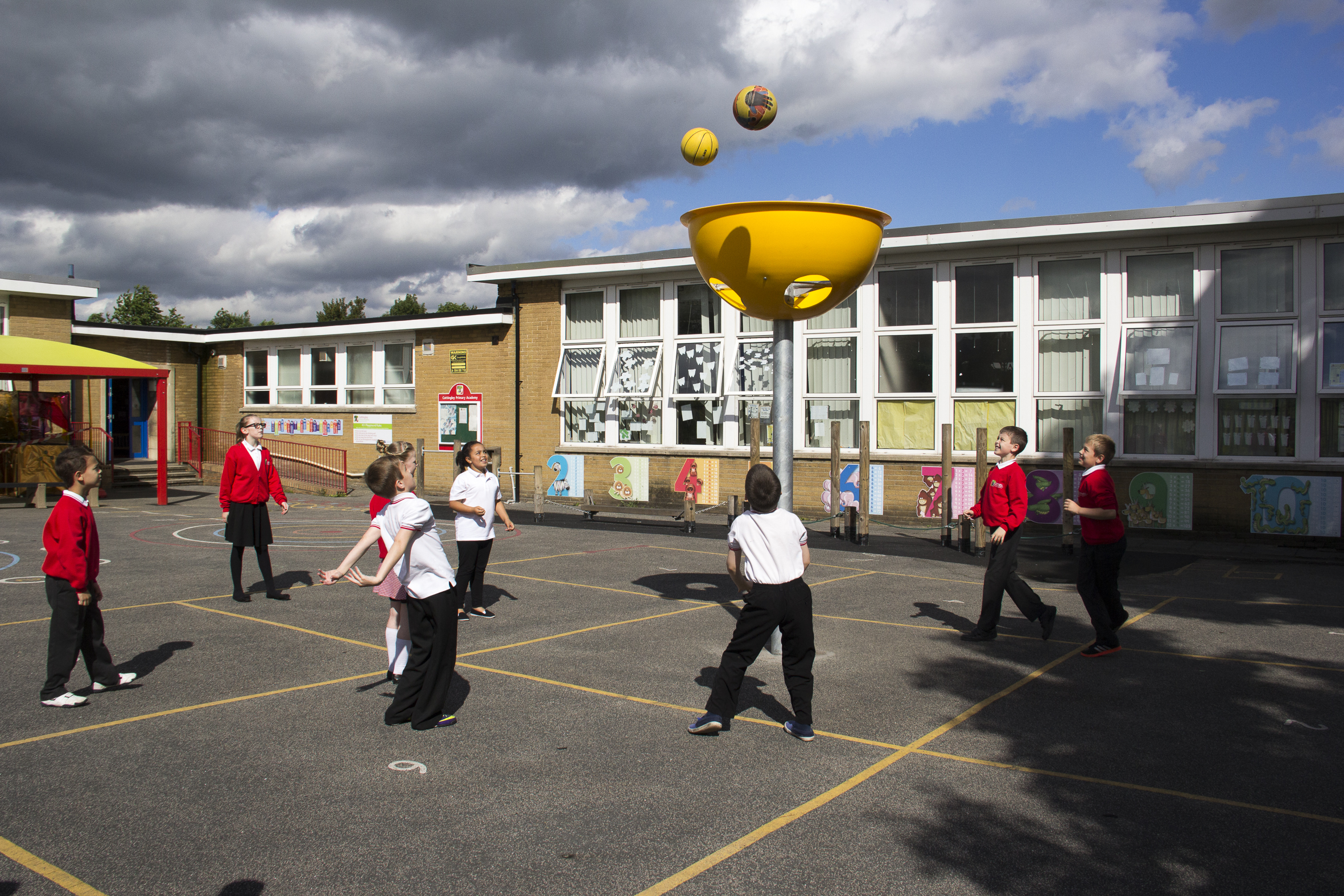 Children play a game in a school playground, aiming balls into a large yellow basket elevated above the ground.