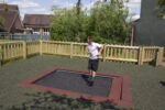 A boy in a school uniform jumps on a sunken trampoline in an outdoor fenced playground area.