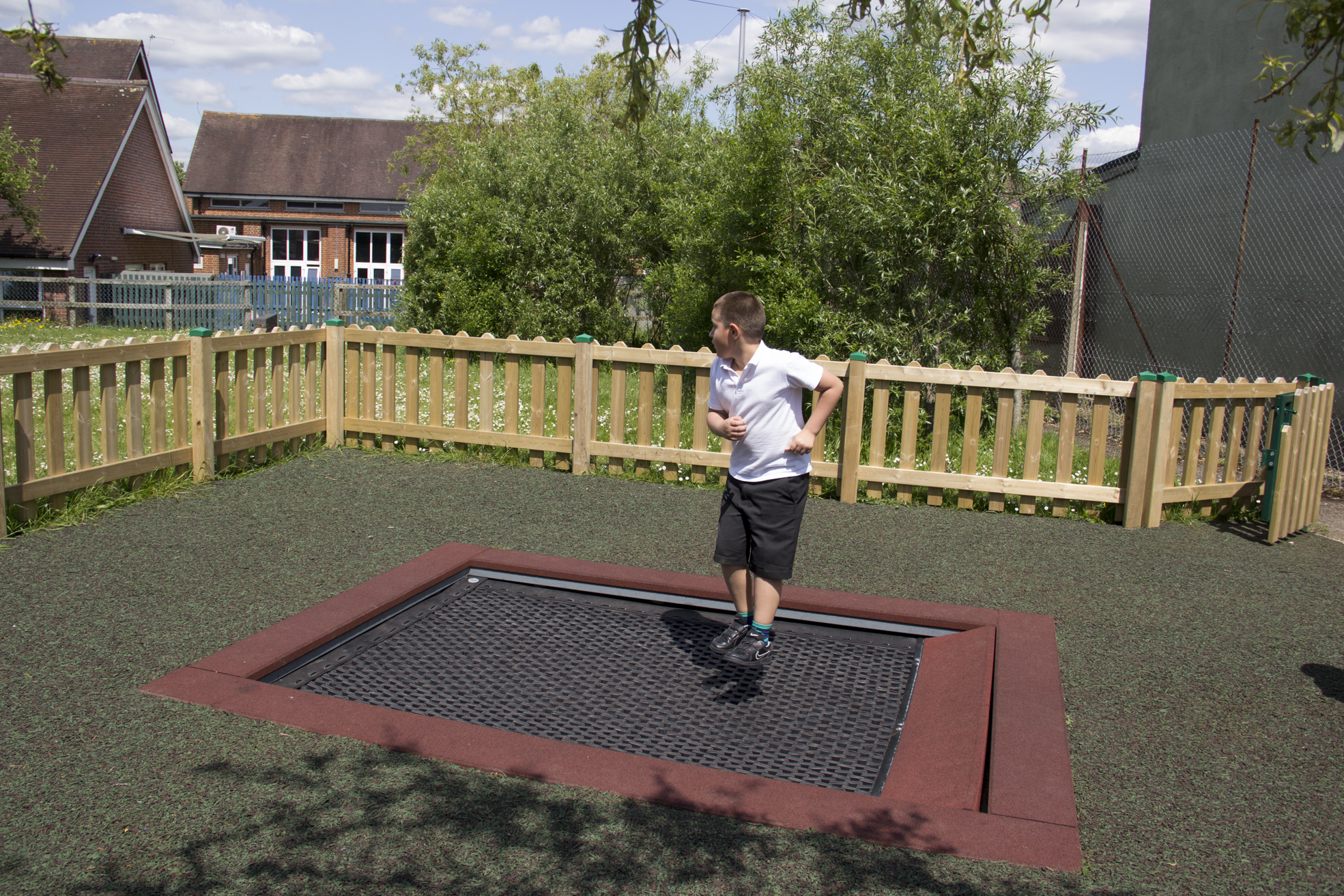 A boy in a school uniform jumps on a sunken trampoline in an outdoor fenced playground area.