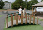 Two girls stand and smile at each other on a small wooden bridge in a playground, with a gazebo, a van, and a picnic table visible in the background.