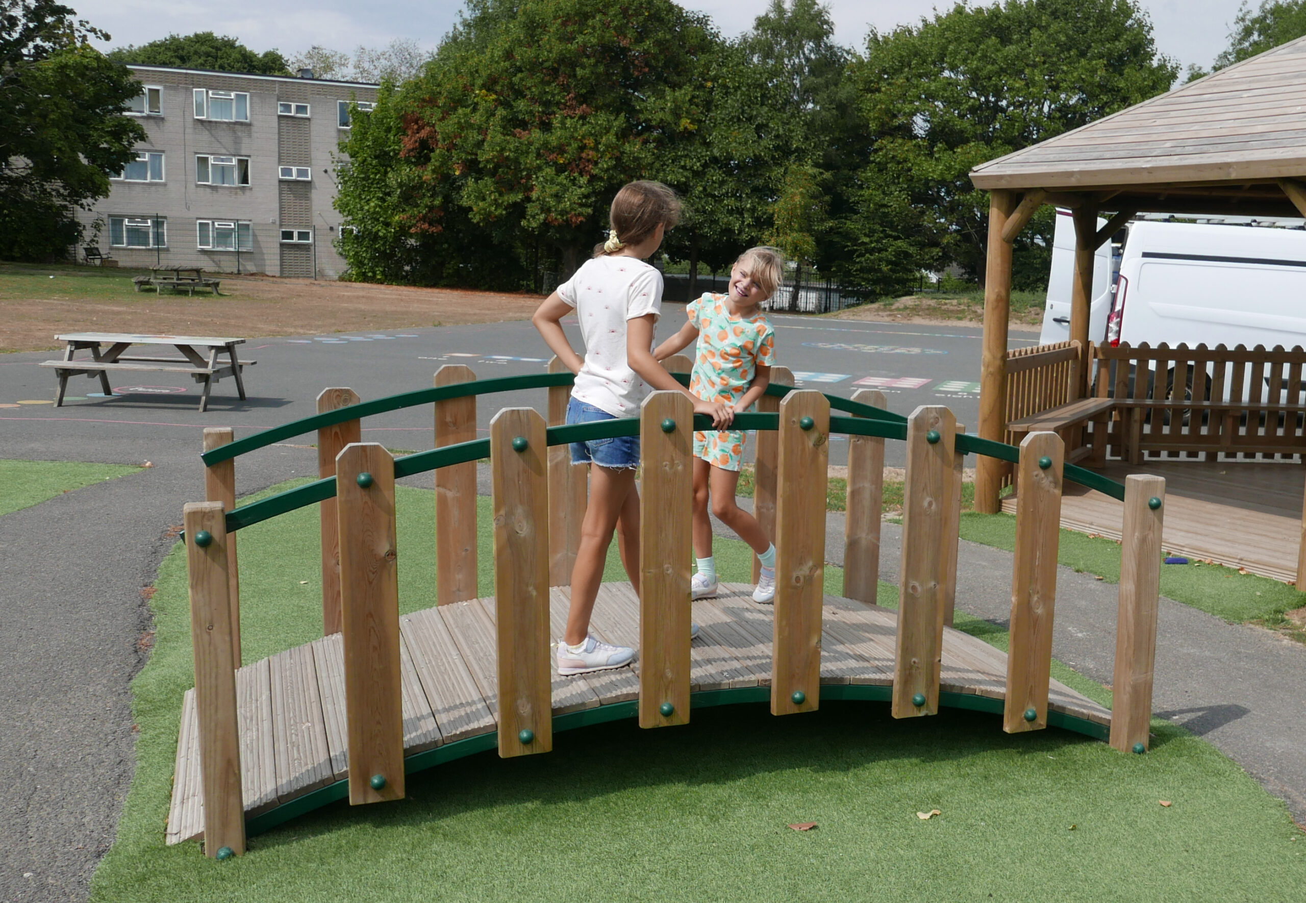Two girls stand and smile at each other on a small wooden bridge in a playground, with a gazebo, a van, and a picnic table visible in the background.