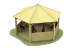 A hexagonal wooden gazebo with a pitched roof, open sides, benches, and four children in red shirts inside, sitting and playing.