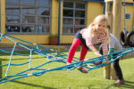 A young girl in red tights climbs on a blue rope net at a playground, whilst another child stands nearby.