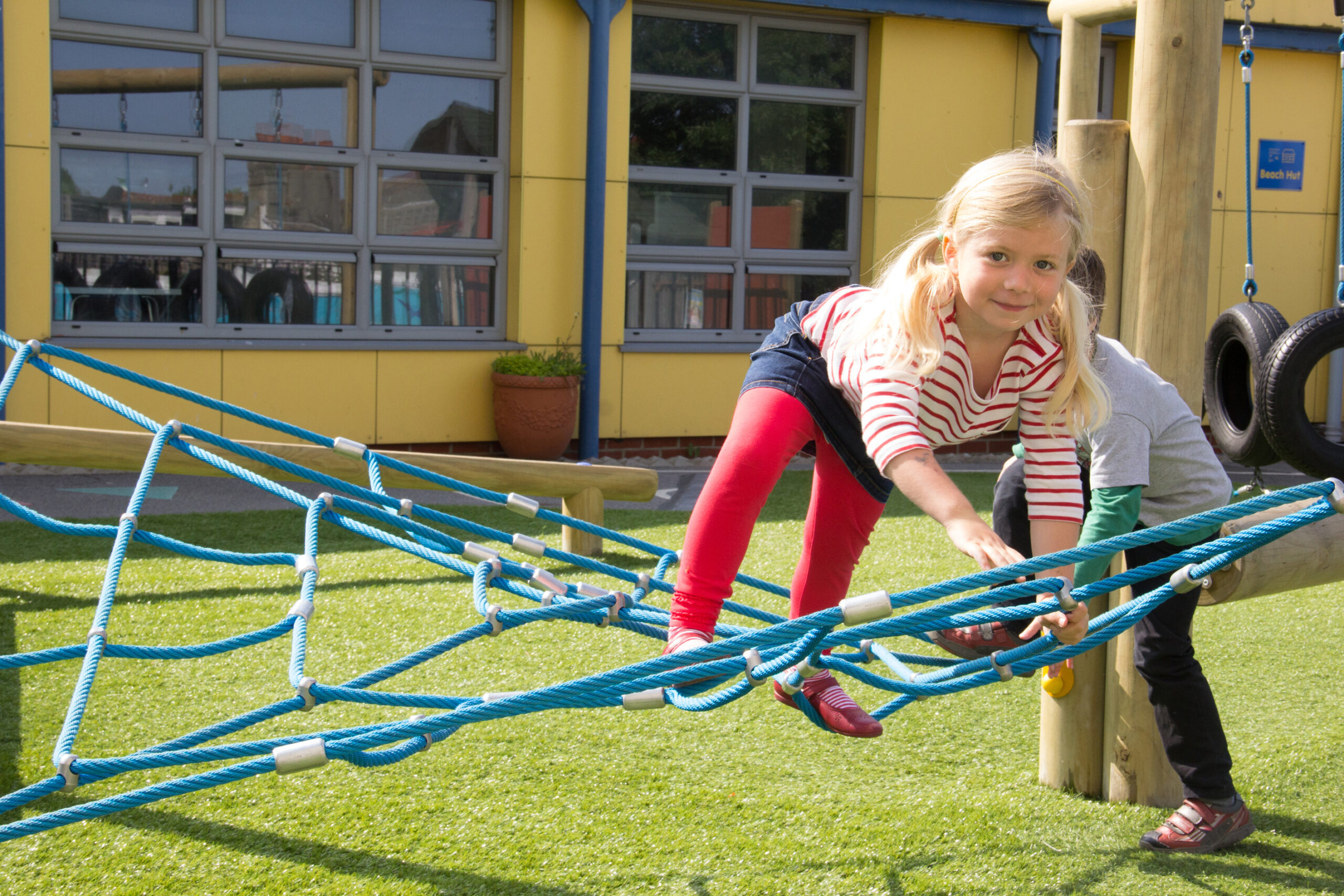 A young girl in red tights climbs on a blue rope net at a playground, whilst another child stands nearby.