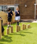 Two young children in school uniforms balance and walk across a row of wooden stepping posts on a grassy play area.