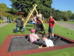 Children, including two in wheelchairs, play on and around a trampoline in an outdoor playground with a supervising adult nearby.