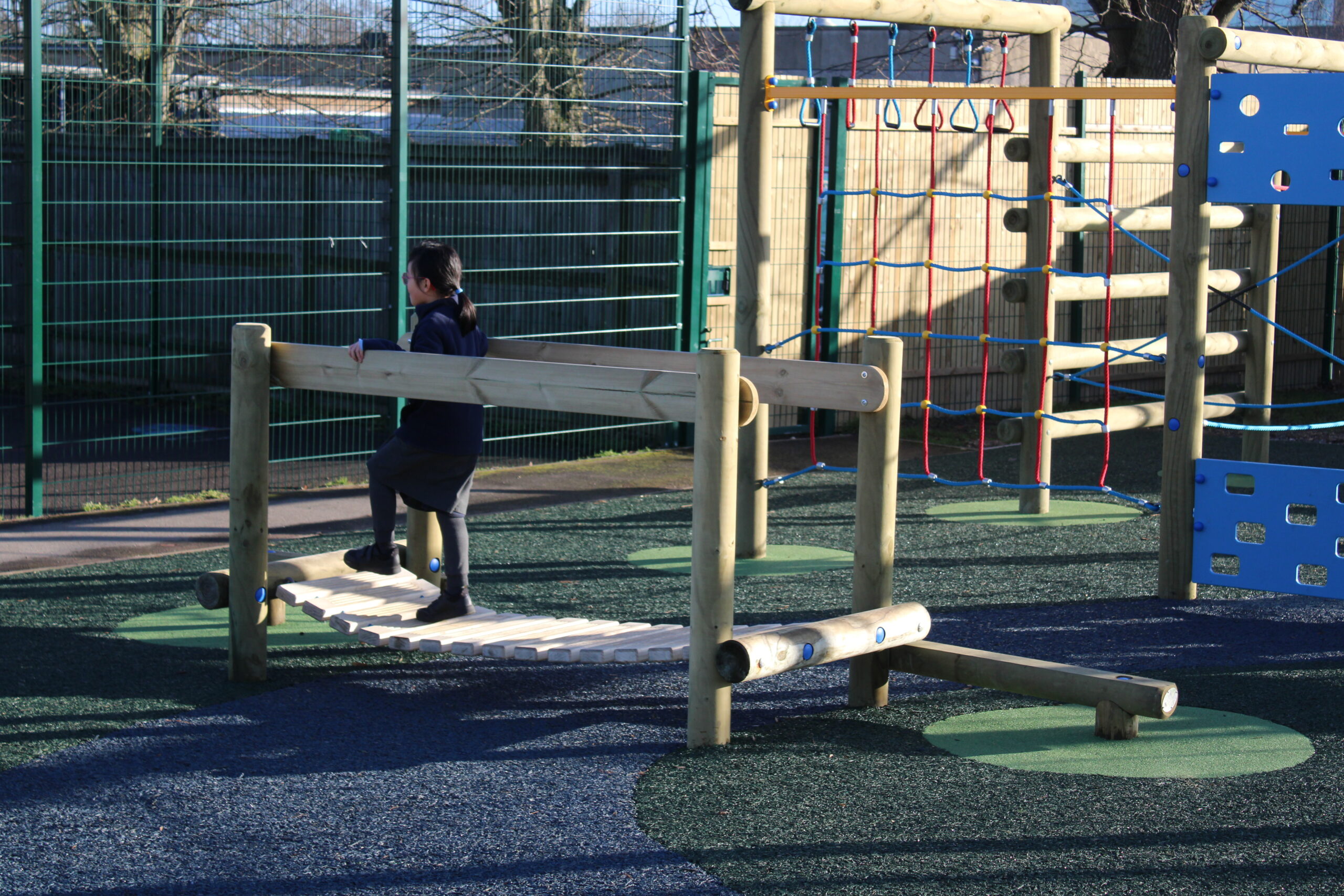 A child walks on a wooden balance beam structure at an outdoor playground with artificial grass and climbing equipment.
