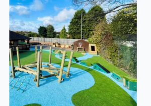 Outdoor playground featuring small playground ideas like climbing equipment, artificial grass, a blue safety surface, and various play structures, all surrounded by trees and a fence.