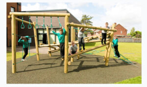 Six children in school uniforms play on a wooden climbing frame, showcasing small playground ideas outdoors, with buildings and grass visible in the background.