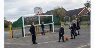 Six children in school uniforms play with a basketball on an outdoor court near a basketball hoop, showcasing small playground ideas with houses and trees in the background.