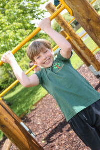 A child smiles whilst hanging from yellow monkey bars at an outdoor playground on a sunny day, showing how active play can improve pupil mental health.