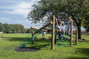 Several children play on a wooden climbing structure with rope nets in a grassy park on a sunny day, enjoying activities that can help improve pupil mental health.