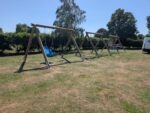 Four wooden swing sets stand on a grassy field under a clear sky, featuring an Inclusive Seat and a Cradle Seat among standard swings; trees and a white van are in the background.