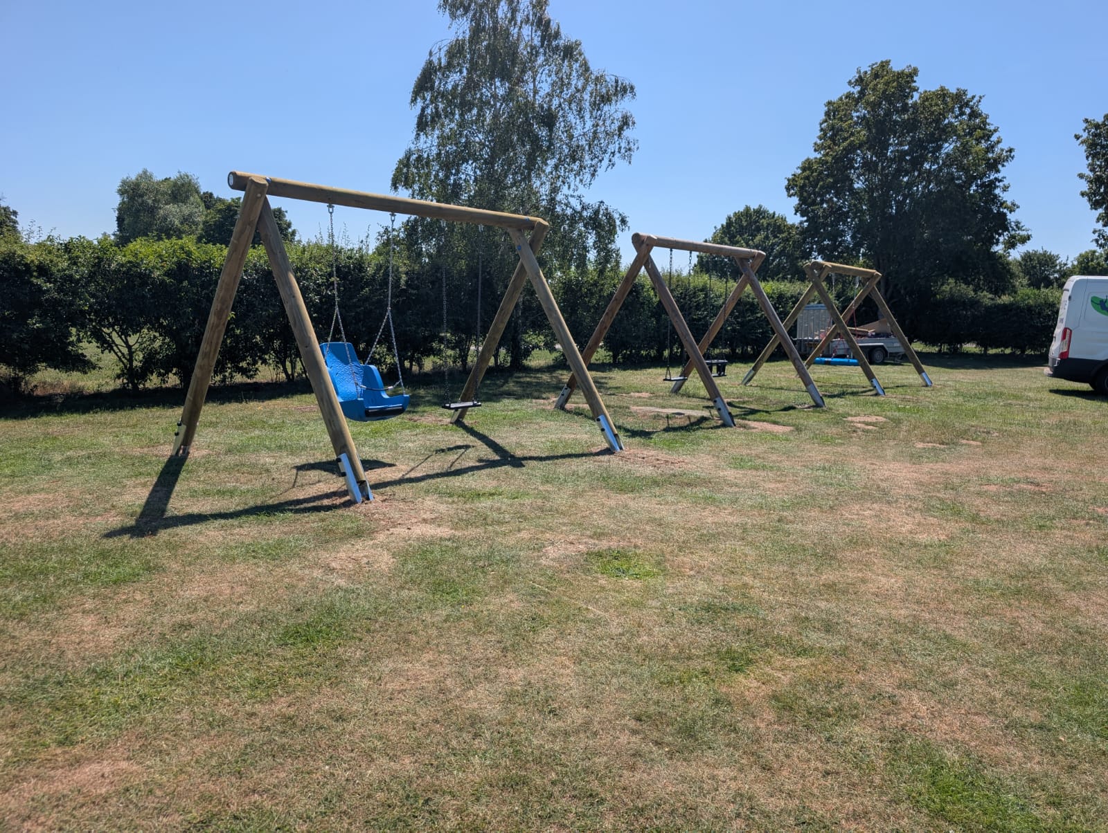 Four wooden swing sets stand on a grassy field under a clear sky, featuring an Inclusive Seat and a Cradle Seat among standard swings; trees and a white van are in the background.