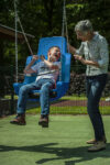 An older woman pushes a smiling boy in a blue inclusive seat swing at an outdoor playground on a sunny day.