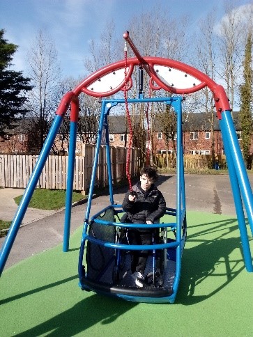 A boy sits in a James Rennie wheelchair-accessible swing at a playground on a sunny day, surrounded by trees and residential houses.