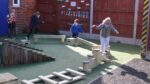Four young children balance and walk on wooden play structures in an outdoor playground area with a fence and numbers chart in the background.