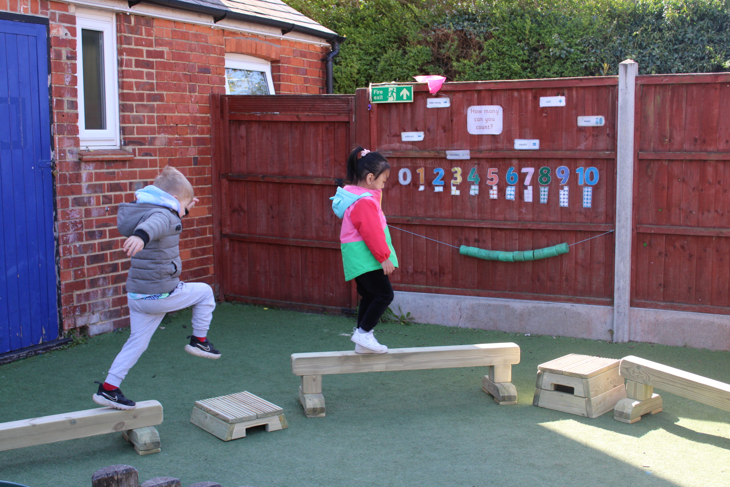 Two children are playing and balancing on wooden beams in an outdoor playground area next to a red fence with numbers displayed on it.