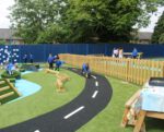 Children play in an outdoor playground with artificial grass, a mini road path, climbing structures, and a wooden fence, surrounded by trees and blue fencing.
