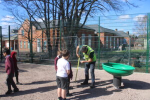 A man in a high-visibility vest measures the ground near a green playground roundabout during a Term Time Installation as children stand nearby on a school playground.