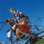 Children climbing on a rope play structure outdoors under a clear blue sky.