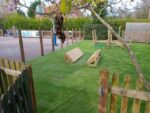 Fenced playground area with artificial grass, wooden climbing equipment including a Clamber Crossing, and adjacent paved play space; trees and fencing line the perimeter.