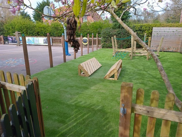 Fenced playground area with artificial grass, wooden climbing equipment including a Clamber Crossing, and adjacent paved play space; trees and fencing line the perimeter.