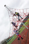 Three children climb on a red rope climbing frame at an outdoor playground on a cloudy day.