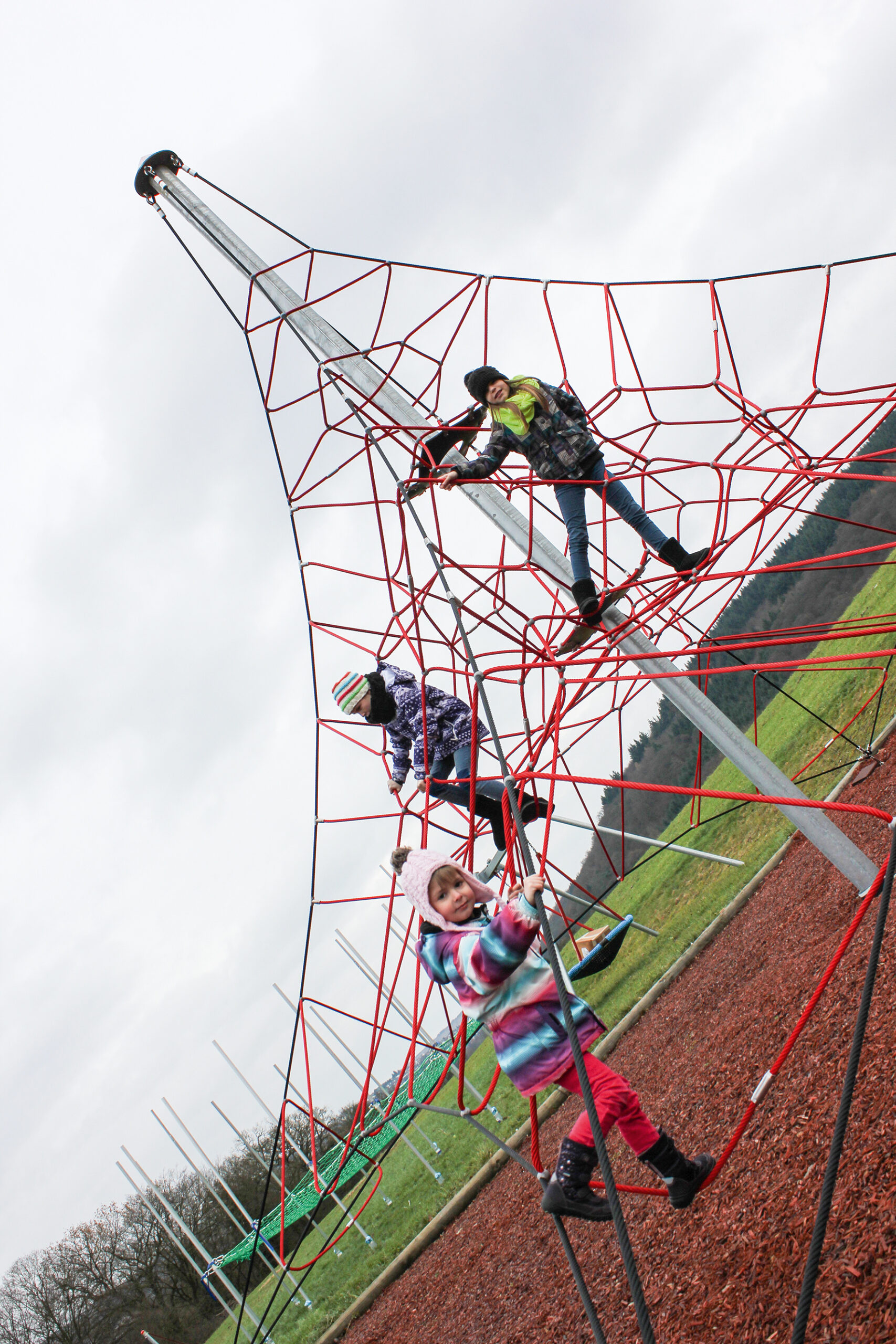 Three children climb on a red rope climbing frame at an outdoor playground on a cloudy day.