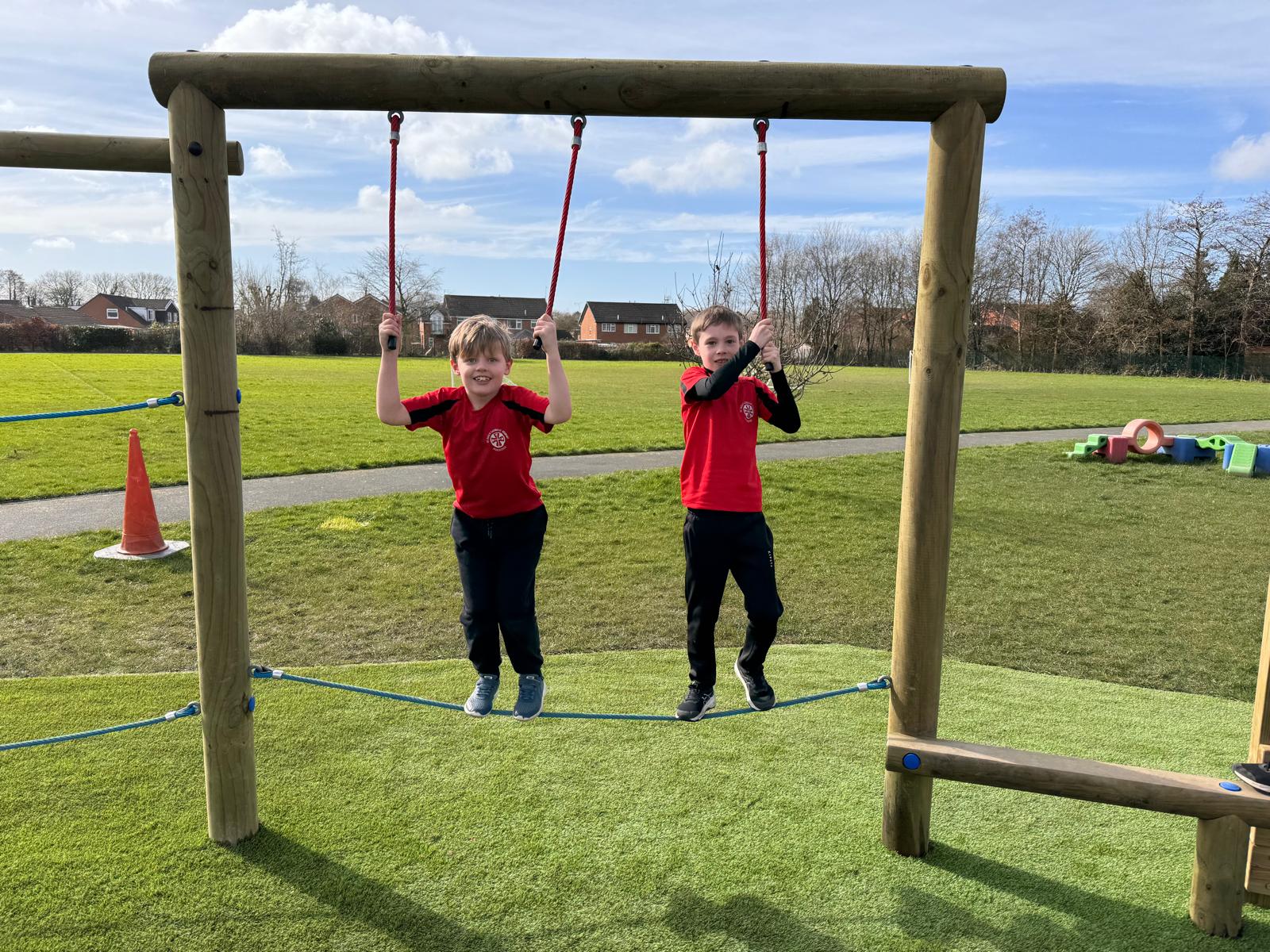Two boys in red shirts climb a wooden playground structure with ropes at St Paul’s, with grass, a path, and houses in the background under a partly cloudy sky.