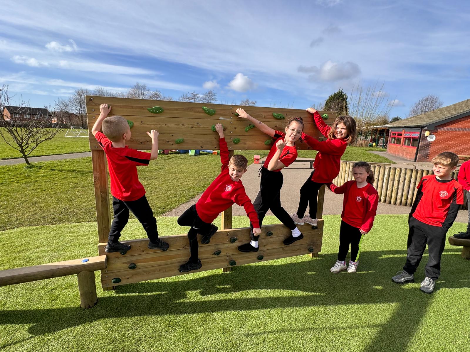 Six children in red uniforms from St Paul’s climb and play on an outdoor wooden climbing wall at a school playground on a sunny day.