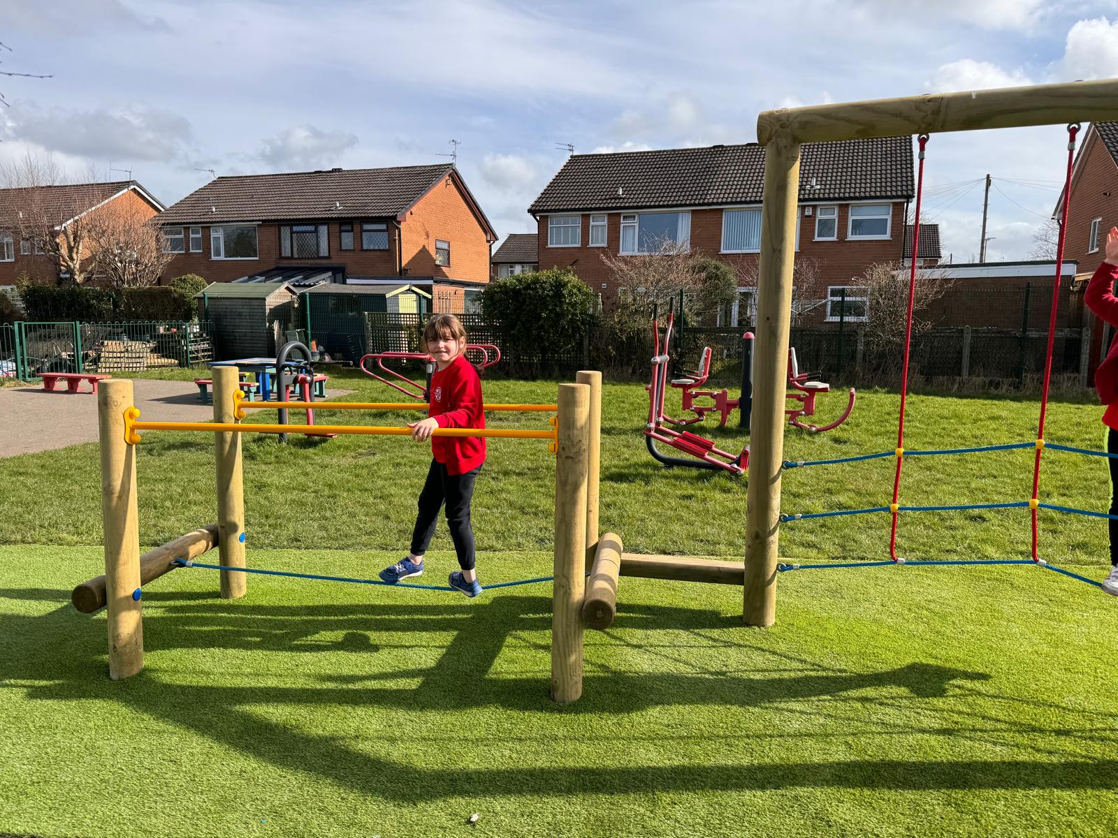 A child in a red jumper balances on a playground obstacle made of ropes and wooden beams on artificial grass in St Pauls, with houses visible in the background.