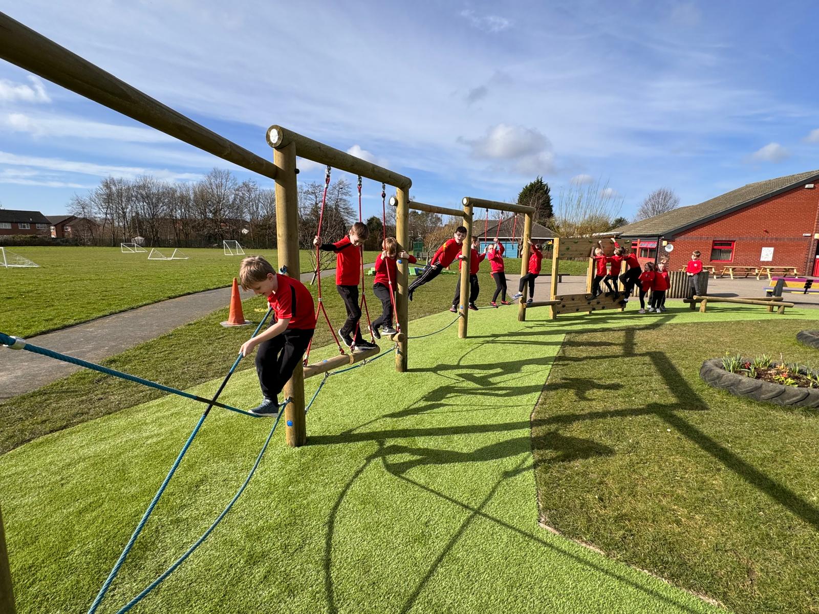 Children in red uniforms play on a wooden balance structure with ropes at St Paul’s playground, with a grassy field and a red brick building in the background.