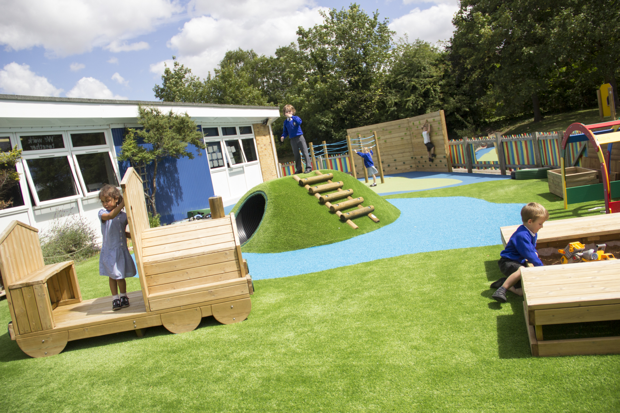 Children play on various playground equipment, including a wooden train, a climbing mound, and a sandpit, in a grassy outdoor area near a school building that feels like a second home.