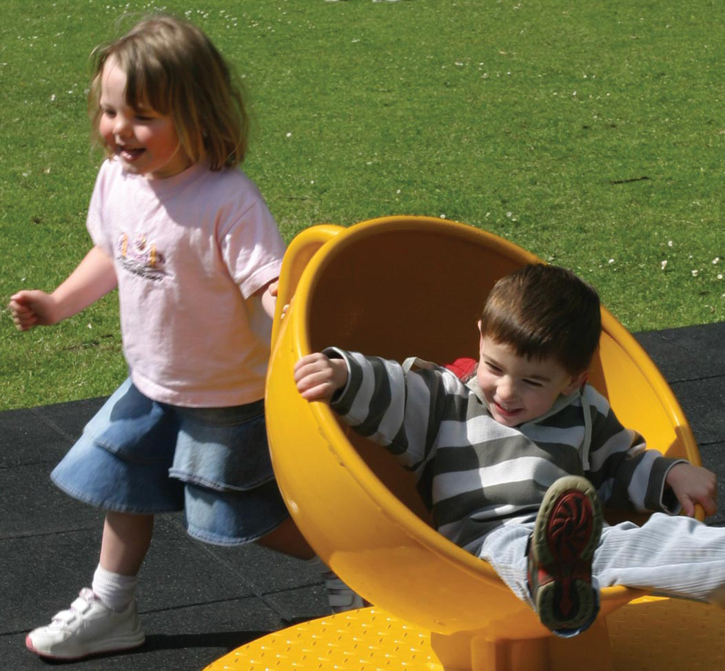 A girl pushes a boy spinning in a yellow Teacup Twister roundabout seat on a sunny day, with grass and rubber playground flooring visible.