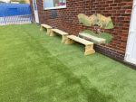 Three wooden benches are placed on artificial green grass against a brick wall with mirror panels.