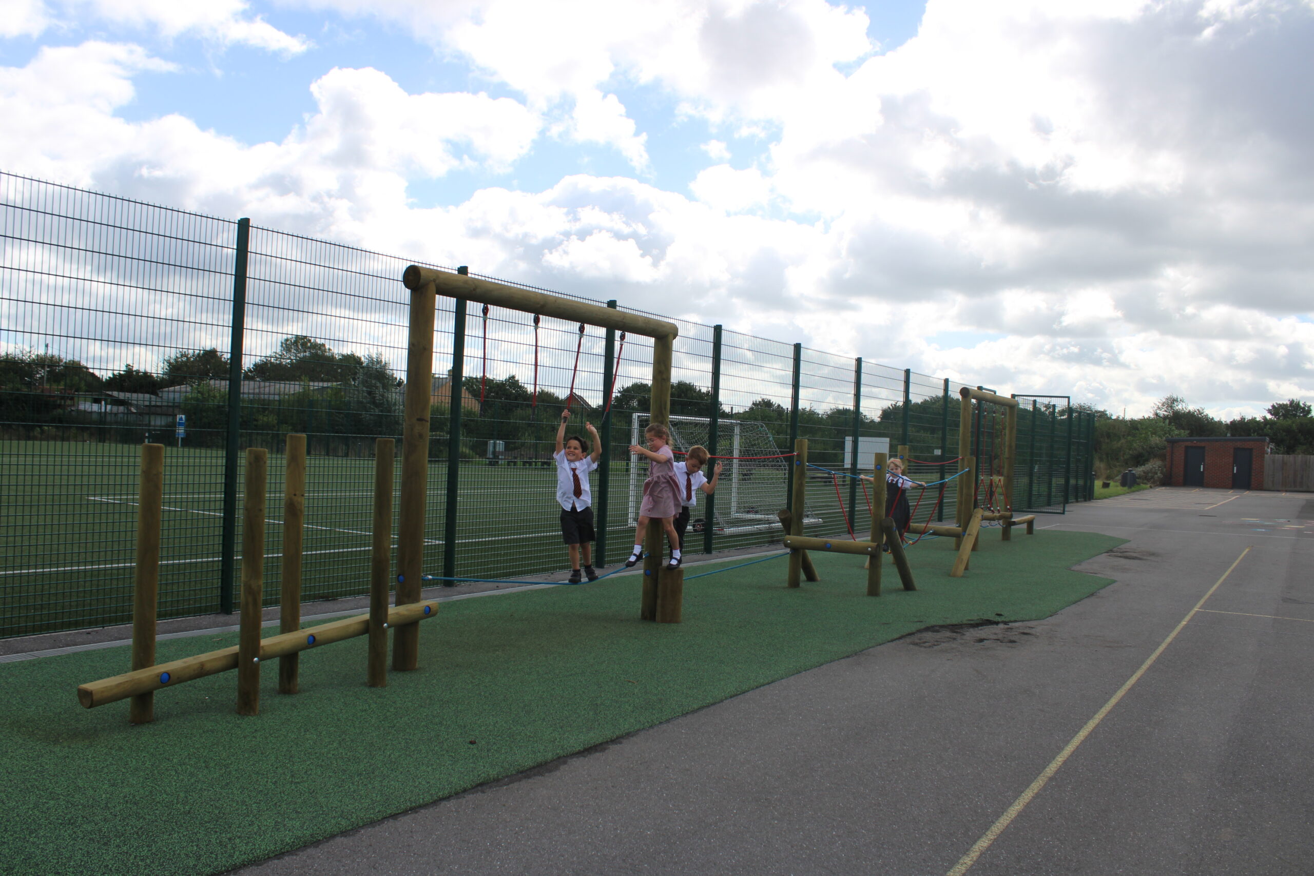 Three children use a wooden climbing frame beside the fenced sports pitch at Nerrols Primary School on a cloudy day.
