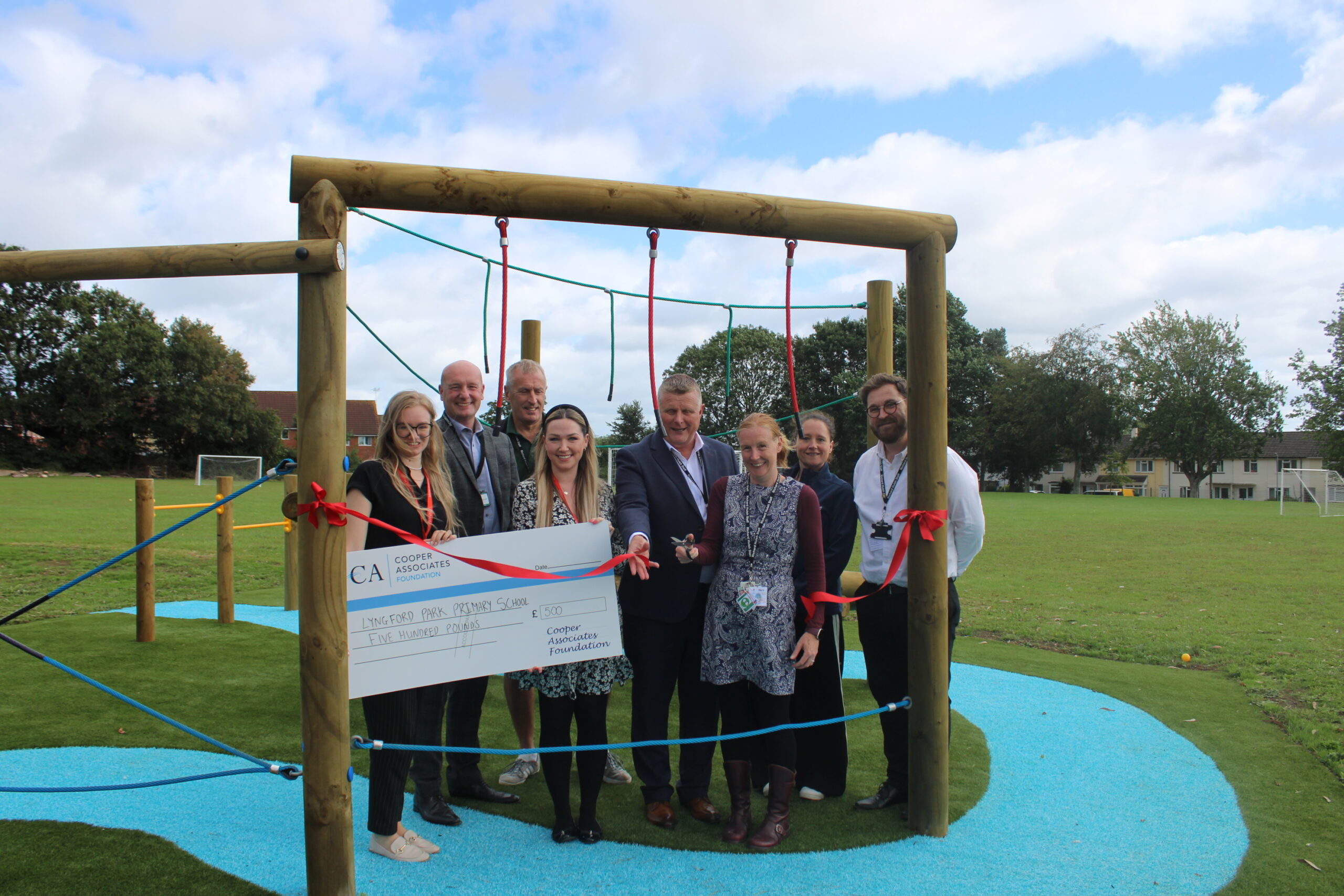 A group of eight adults stands in front of a rope climbing structure at Lyngford Park, holding a large cheque on a grassy field, with ribbon and playground equipment visible.