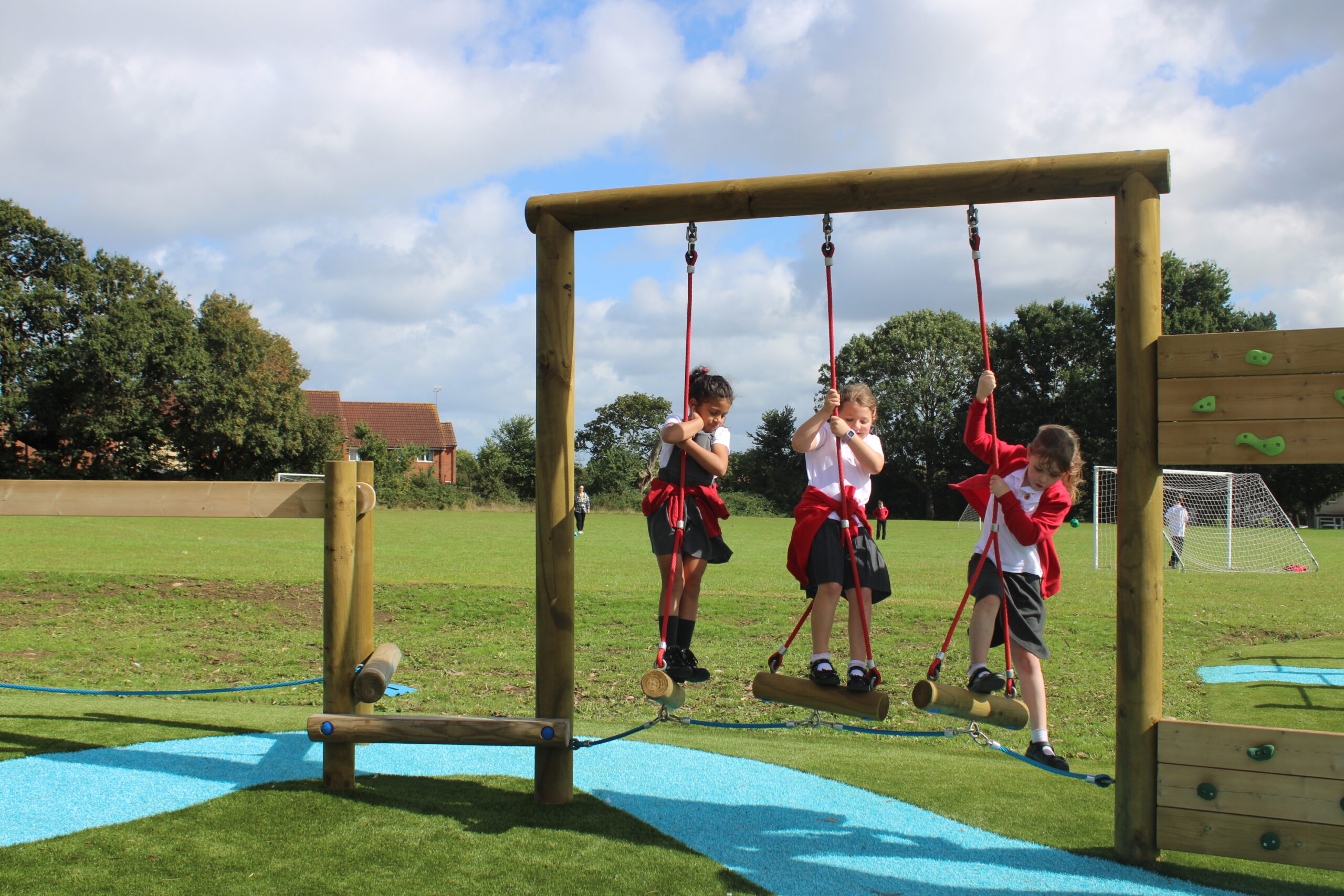 Three children in school uniforms play on a wooden swing obstacle at Lyngford Park playground, with a grassy field and football goals in the background.