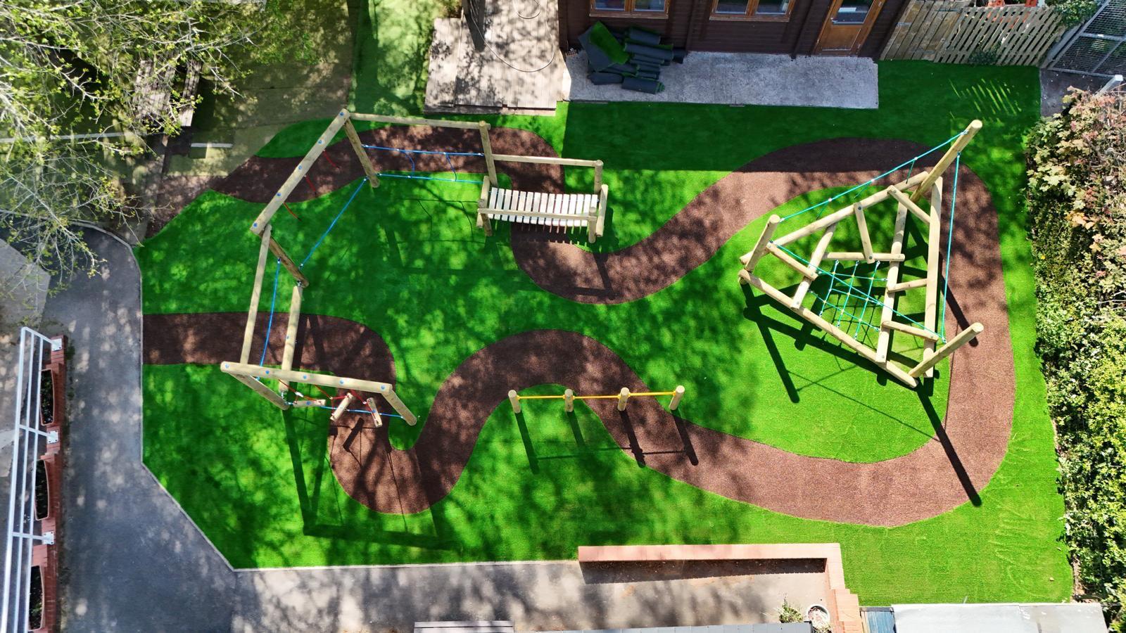 Aerial view of a playground with artificial grass, swings, a climbing structure, and a winding brown path, surrounded by trees and a building.