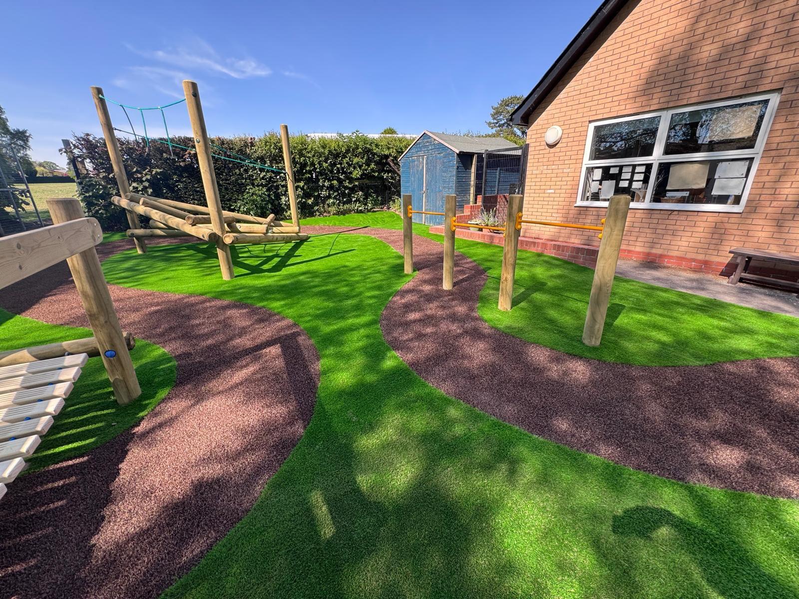 Outdoor playground area with artificial grass, balance beams, climbing structure, and a brick building in the background under a clear blue sky.