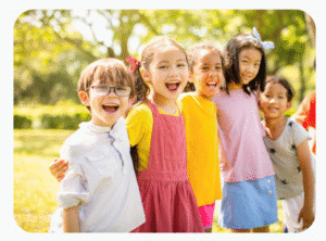 Five children stand in a row outdoors, smiling and laughing on a sunny day with trees and grass in the background.