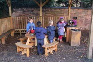 Five young children play together outdoors on wooden benches and tables, with two exploring a mud kitchen set under a wooden shelter. This setting encourages EYFS Communication and Language skills, surrounded by leaves and a brick wall.