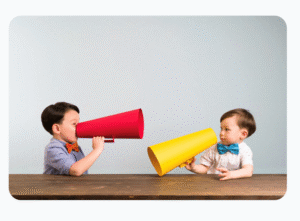 Two young boys sit at a table facing each other, holding large red and yellow megaphones, appearing to speak through them.