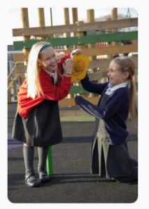 Two young girls in school uniforms play with a yellow talking device on a playground, smiling and interacting as they develop EYFS Communication and Language skills together.