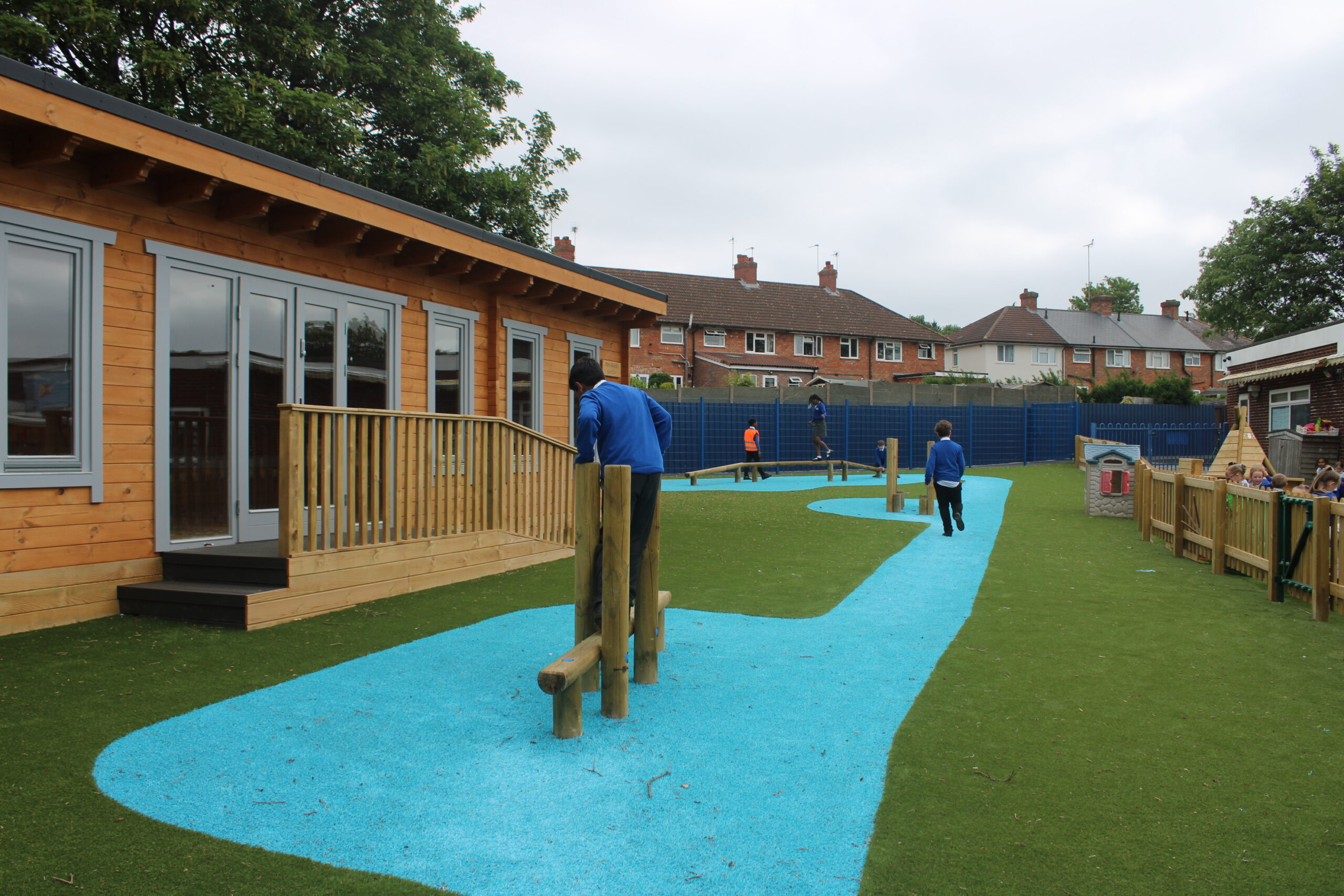 Children play outside on artificial grass with a winding blue path near a wooden building, surrounded by houses and trees on a cloudy day.