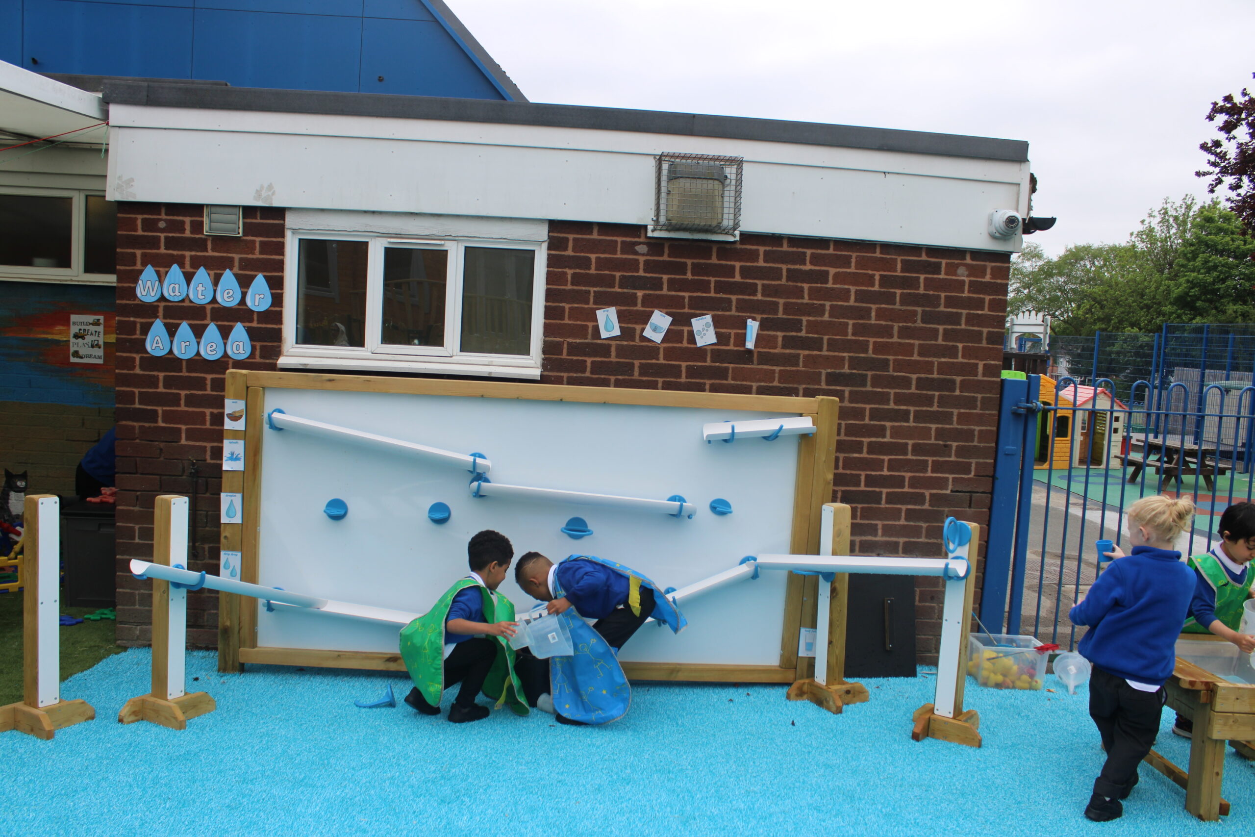 Three young children play with an outdoor water wall and pipes on a blue mat beside a brick building at a school or daycare.