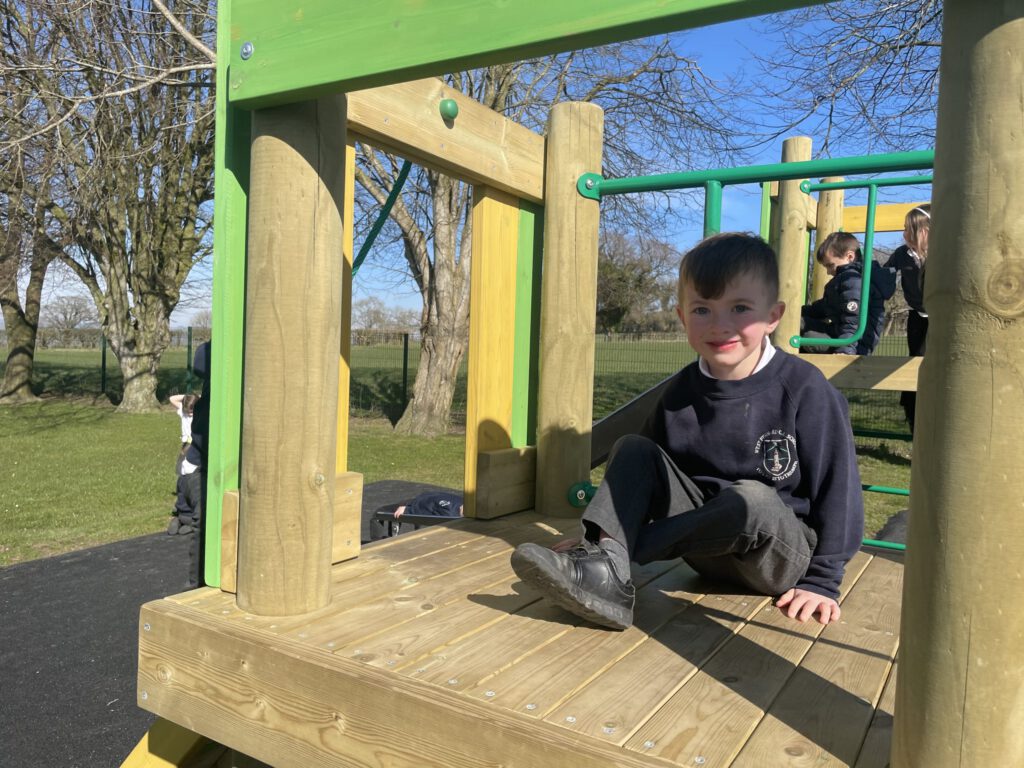 A child sits on a wooden playground structure in West Pennard, ready to slide. Other children play in the background as sunlight casts shadows across the area. Tall trees sway gently, adding a serene backdrop to the lively scene.