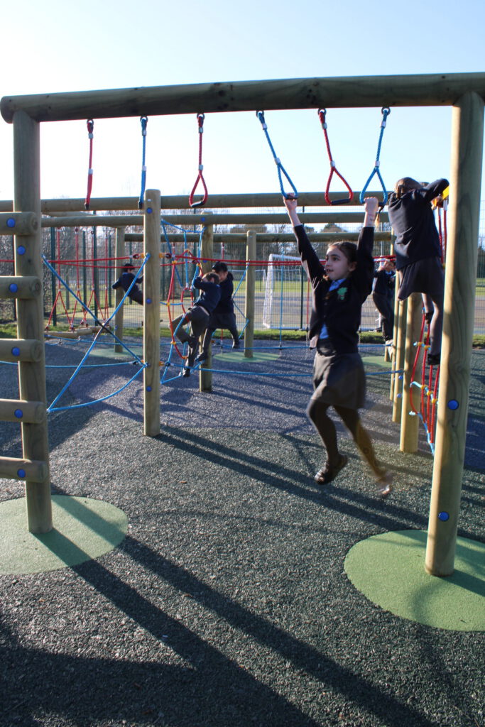 Children in school uniforms from Westende Junior play on a wooden climbing frame and monkey bars at an outdoor playground on a sunny day.