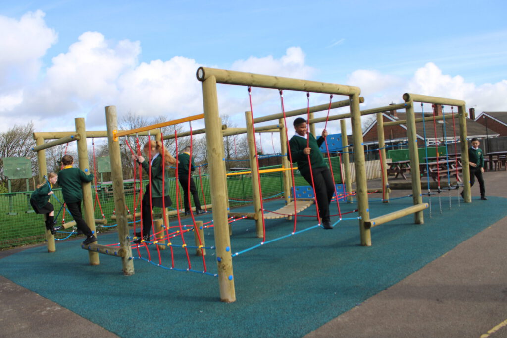 Children in Sutherland Primary Academy uniforms play on a wooden climbing frame with ropes at an outdoor playground on a sunny day.